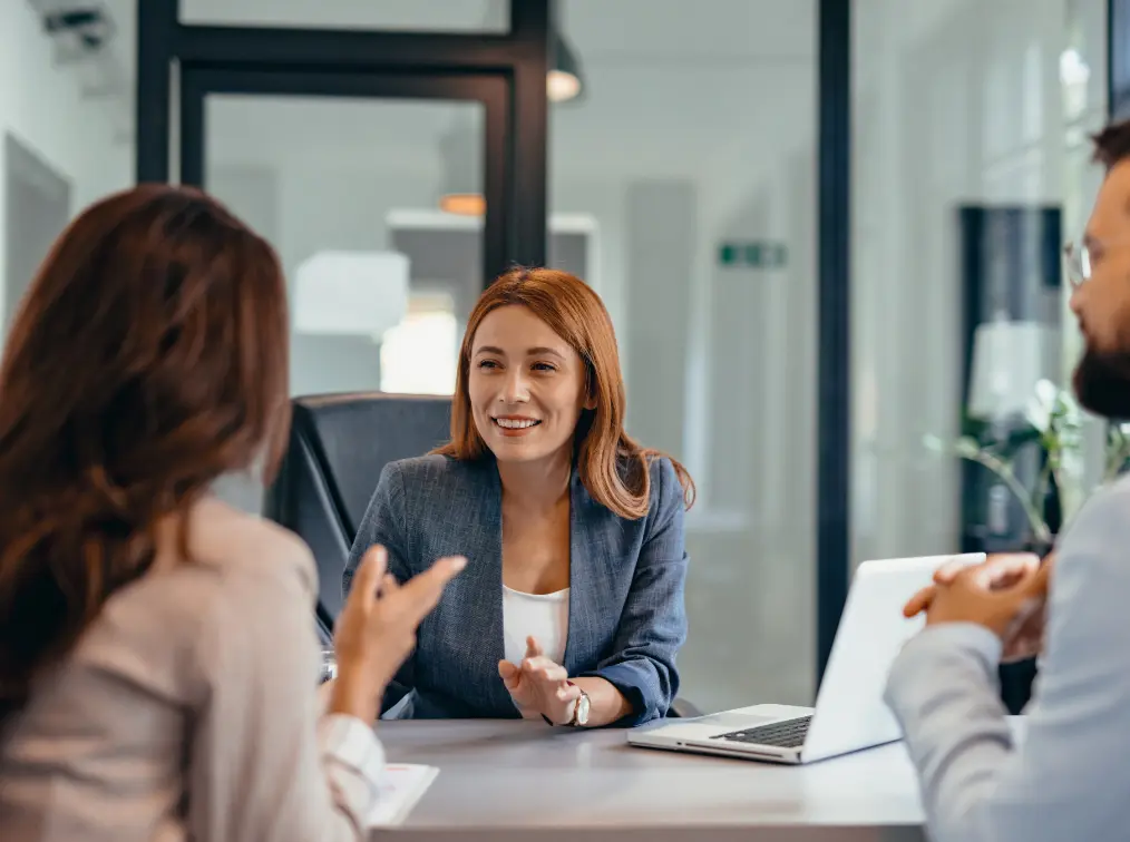 Three people sit at a table in an office setting. A woman in a blazer smiles while talking to a woman and man sitting across from her. A laptop is open on the table.