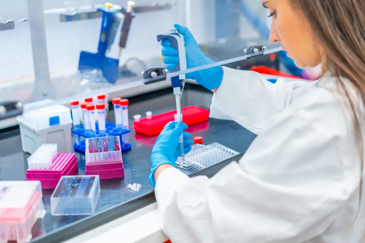 A scientist in a lab coat and gloves uses a pipette to transfer liquid into a microplate, surrounded by vials and lab equipment.