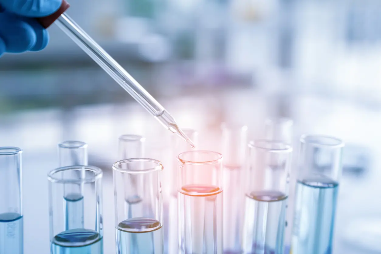 Close-up of a pipette above test tubes containing blue liquid in a laboratory setting.