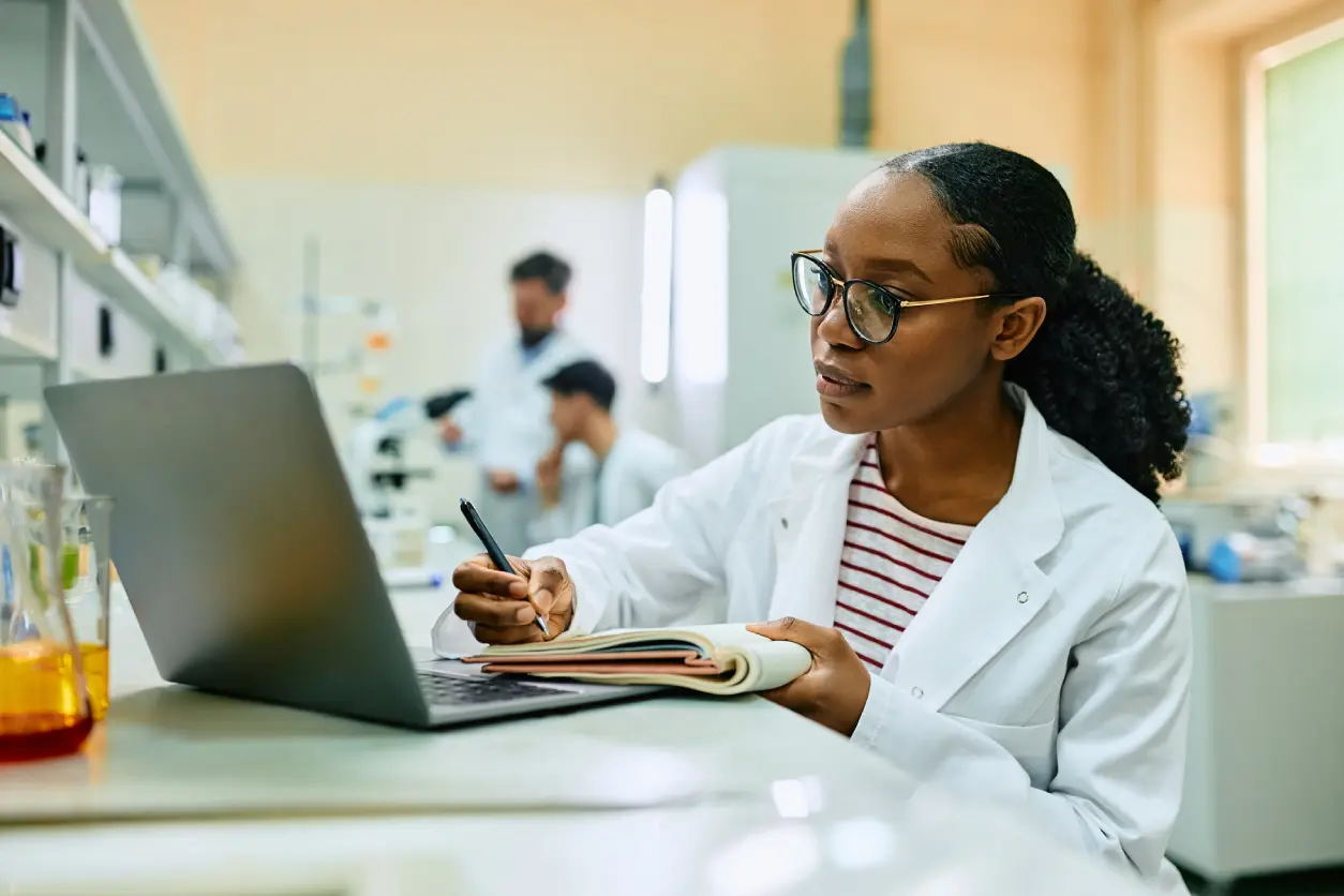 A scientist in a white lab coat writes in a notebook while looking at a laptop. Two other scientists work with microscopes in the background.