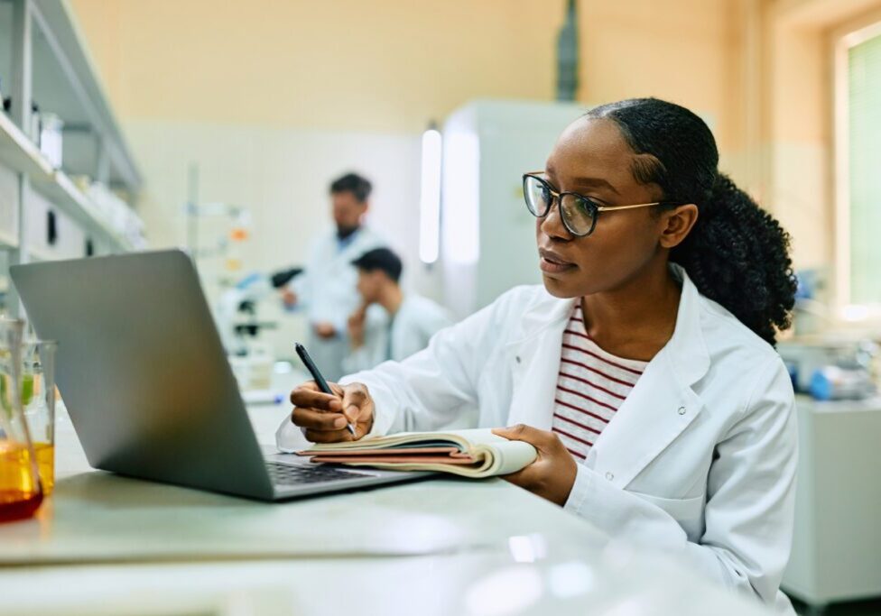 A scientist in a white lab coat writes in a notebook while looking at a laptop. Two other scientists work with microscopes in the background.