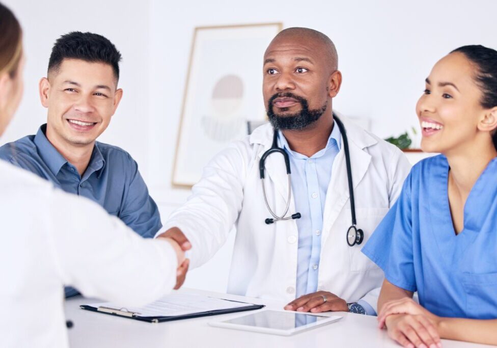 A doctor and nurse sit at a table, shaking hands and smiling with a patient. Medical documents and a tablet are on the table.
